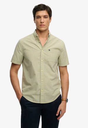 Jeune homme portant une chemise à manches courtes à carreaux jaune avec un petit logo sur la poitrine, un pantalon sombre et une montre-bracelet avec un bracelet en cuir marron.