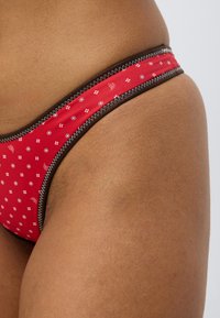 Κόκκινο κάτω μέρος μπικίνι με λευκά ανθικά και γεωμετρικά σχέδια, με επένδυση από σκουρό καφέ ύφασμα, που παρουσιάζει ομαλή υφή και μέτριο ύψος στη μέση.