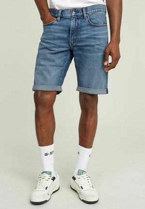 Homme portant un short en denim bleu roulé, des baskets blanches et des chaussettes blanches montantes avec des inscriptions noires sur un fond uni.