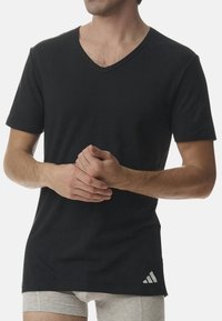 Svart t-shirt med kort ärm och V-hals, tillverkad av mjukt tyg, med en vit Adidas-logotyp på nedre vänstra sidan. Kroppsnära passform.