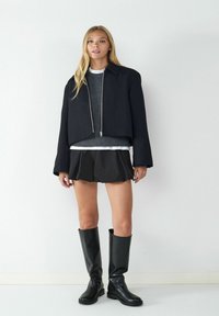 Kurzes schwarzes Jackett mit silbernem Reißverschluss, grauer Pullover darunter, schwarze plissierte Shorts und hohe schwarze Lederstiefel. Schlichter Hintergrund.