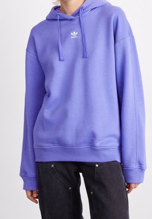 Osoba ubrana w fioletową bluzę z kapturem Adidas z sznurkami i czarne dżinsy, stojąca na jednolitym jasnym tle.