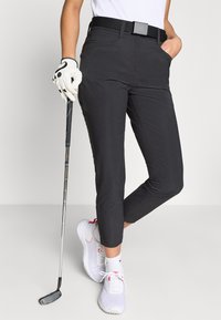 Μαύρο παντελόνι γκολφ με εφαρμοστή σχεδίαση, διαθέτει θηλιές για ζώνη, τσέπες και κοντό μήκος. Συνδυάζεται με λευκά και ροζ παπούτσια γκολφ.