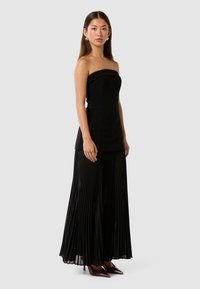 Svart strapless kjole med en tettsittende bodice og en plisserte skjørt. Stoffet har en glatt tekstur, komplementert med spisse hæler.