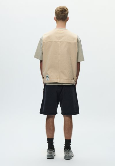 Mężczyzna stojący tyłem, ubrany w beżową kamizelkę bez rękawów na szarej koszulce, czarne szorty, czarne skarpetki i szare tenisówki na jednolitym tle.