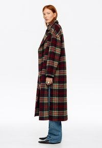 Mujer joven vistiendo un abrigo largo de cuadros rojos y verdes, con vaqueros azules y zapatos negros puntiagudos, de pie de perfil sobre un fondo blanco.