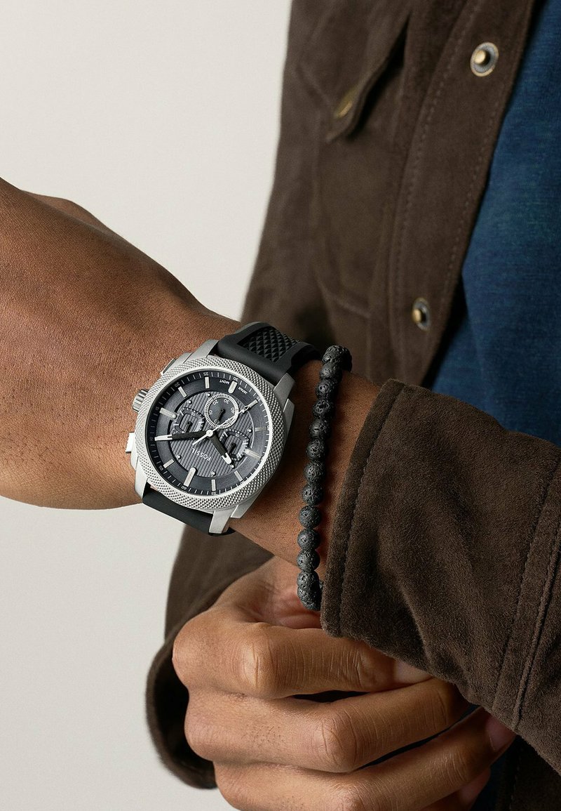 Montre avec bracelet en silicone noir, boîtier texturé en argent, cadran gris avec trois sous-cadrans et index lumineux ; portée sur un poignet orné d'un bracelet en perles.