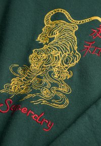 Groen stof met een geel geborduurde tijger en rode geborduurde Japanse tekens en de merknaam "Superdry".