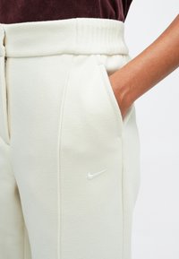 Šviesių kreminių jogger tipo kelnių pagamintų iš švelnios, tekstūruotos medžiagos. Yra elastinė juosmens juosta, šoninės kišenės ir balta Nike logotipas ant šlaunies.
