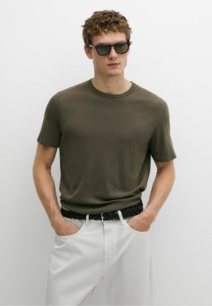 Giovane uomo con capelli ricci indossa occhiali da sole neri, maglietta verde oliva, pantaloni bianchi e cintura intrecciata nera con le mani nelle tasche.
