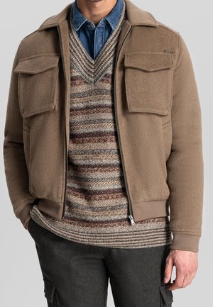 Mand iført en beige lynlåstrøje over en stribet strikket sweater og blå denimskjorte, kombineret med mørke cargobukser, hånd i lommen.