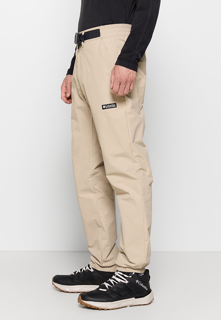 Pantalons imperméables beiges avec taille élastique, ceinture réglable et chevilles resserrées, présentant un patch logo Columbia sur la cuisse.