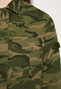 Veste camouflage verte et marron avec une poche poitrine à bouton et un écusson sur la manche, vue de la zone avant de l'épaule.