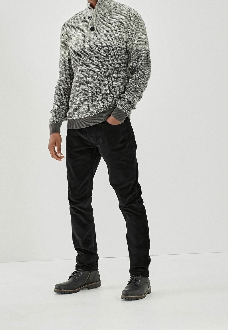 Gebreide grijze en witte trui met een hoge kraag en twee knopen, gecombineerd met zwarte ribfluwelen broek en donkere veterlaarzen.