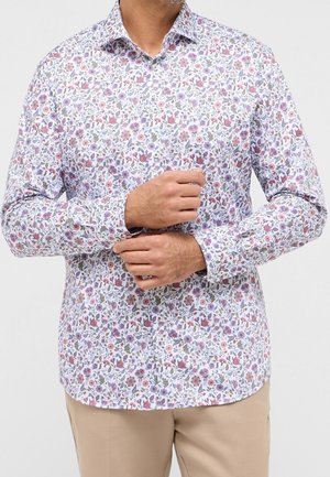 Mann trägt ein weißes, langärmliges Hemd mit Knöpfen und einem floralen Muster in Pink, Lila und Grün sowie beige Hose und richtet die Manschette.