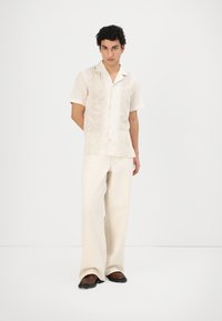 Beige Kurzarmhemd mit strukturierten floralen Stickereien, kombiniert mit hellen, weiten Hosen und braunen Slippern.