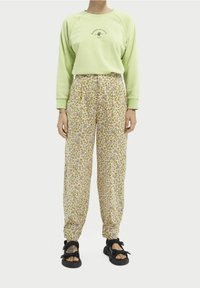 Persoon draagt een lichtgroene trui, hooggetailleerde broek met bloemenpatroon en zwarte open-toe sandalen, staand tegen een witte achtergrond.