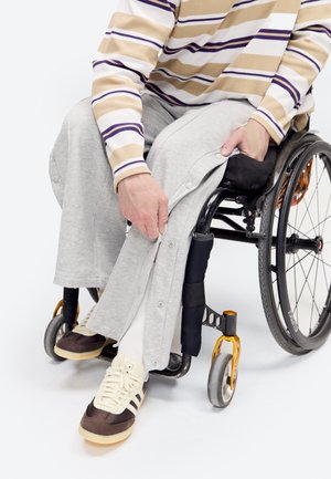 Osoba u invalidskim kolicima podešava sive hlače sa drukerima, nosi prugasti bež, bijeli i ljubičasti džemper s smeđim i krem tenisicama.