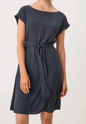 Vrouw draagt een marineblauwe jurk tot over de knieën met korte mouwen, een boothals en een stoffen riem gestrikt rond de taille, staand tegen een neutrale achtergrond.