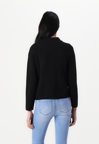 Černý, texturovaný, dlouhý svetr s límcem, nosený s vybledlými modrými úzkými džíny. Oblečení je vidět zezadu.