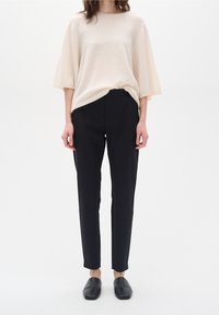 Maglione beige ampio con scollatura larga; abbinato a pantaloni neri su misura che si stringono alla caviglia e scarpe nere slip-on.