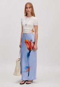 Witte top met korte mouwen en geplooide zijkanten, gecombineerd met een lichtblauwe maxi rok met een grote oranje tulpenprint. Zwarte sandalen en een beige tas.