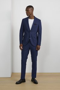 Traje navy con un blazer ajustado y pantalones de corte plano. Llevado sobre una camiseta blanca. Presenta un pañuelo de bolsillo y zapatos negros brillantes.