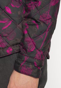 Quiltad jackärm i svart med ett djärvt rosa blommönster. Har kontrasterande sömmar och en slät yta. Texturerad tyg synlig.