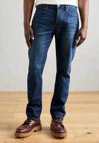 Jeans din denim albastru închis, cu croi drept, care prezintă cusături portocalii și cinci buzunare. Asortați cu pantofi maro cu șireturi și pardoseală din lemn.