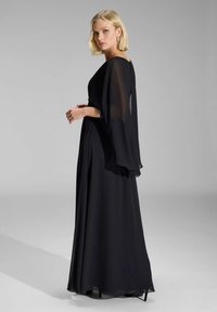 Sort aftenkjole med en svævende chiffonkappe og en slank silhuet, med lange ærmer og en let glans. Enkel og elegant design.