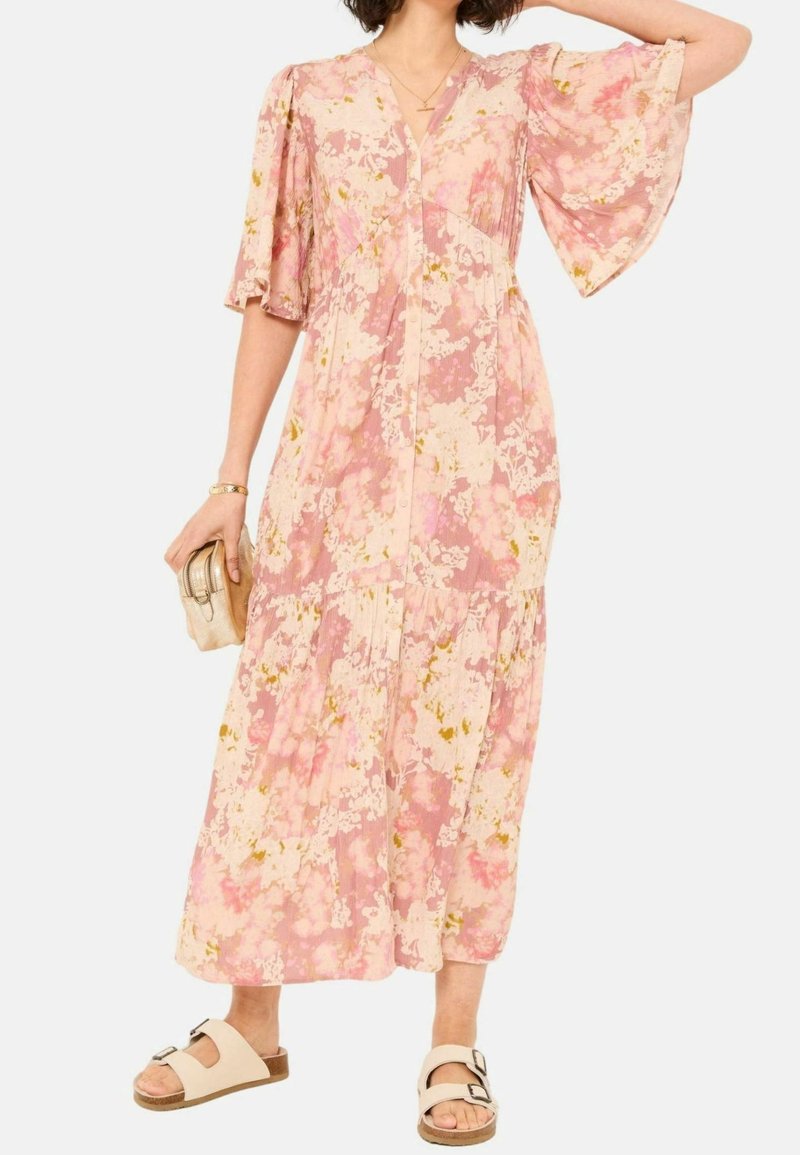 Femme portant une robe midi fleurie rose à larges manches, tenant une petite pochette beige et des sandales beiges avec des boucles.
