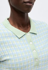 Κοντινό πλάνο ενός ατόμου που φοράει ένα παστέλ πράσινο και μπλε καρό πόλο με τέσσερα λευκά κουμπιά και μυτερό γιακά.