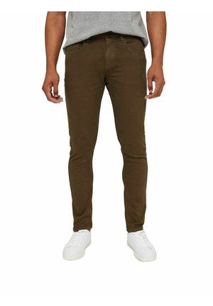 Mand iført brune slim-fit bukser, grå skjorte og hvide sneakers, står foran en ensfarvet hvid baggrund.