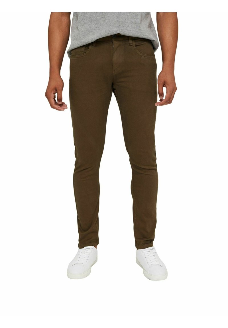 Homme portant un pantalon slim marron, une chemise grise et des baskets blanches, debout devant un fond blanc uni.