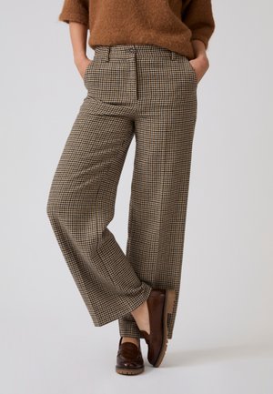 Persoon die bruine geruite wijde broek, een bruine trui en donkerbruine loafers draagt, staand met handen in de zakken en één been gekruist.