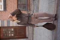Brown faux fur jacket with wide sleeves, brown tailored trousers, and dark brown suede bag. Standing on stone pavement, hair styled back.