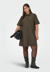 Hnědé šaty s krátkým rukávem a knoflíkovou přední částí, doplněné černou kabelkou a koženými botami po kolena se sponkami.
