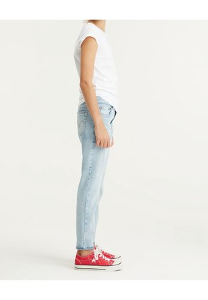 Person iført hvid t-shirt, lyseblå jeans og røde sneakers, der står i profil mod en ensfarvet hvid baggrund.