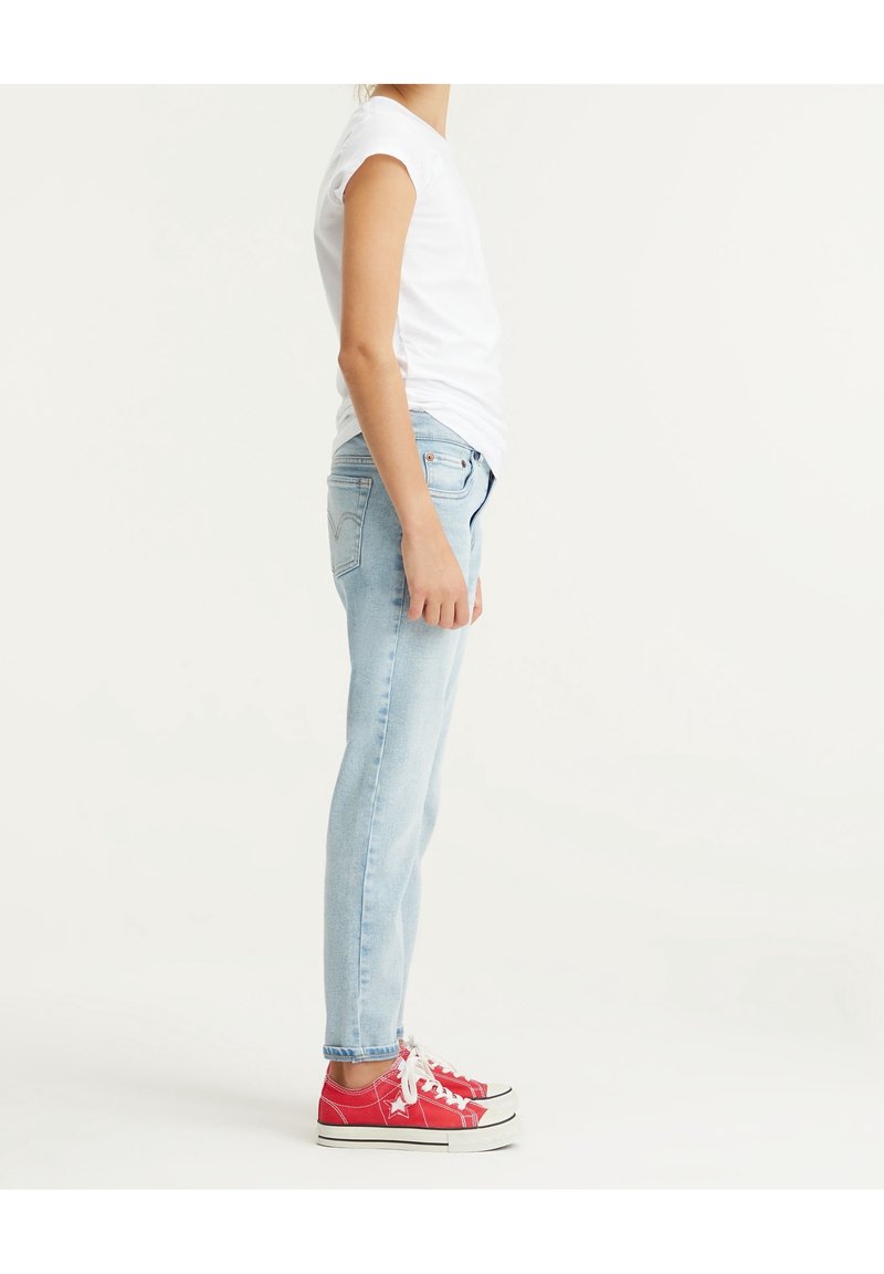 Personne portant un t-shirt blanc, un jean bleu clair et des baskets rouges, debout de profil sur un fond blanc uni.