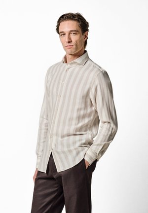 Mand iført en beige og hvid vertikalt stribet skjorte med knapper og mørkebrune bukser, stående med den ene hånd i lommen mod en hvid baggrund.