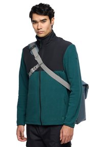 Giacca in pile con parte superiore nera e parte inferiore teal, tasche con zip, colletto alto, indossata con una tracolla grigia e una borsa.