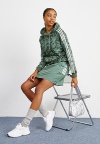 Zöld mintás kapucnis pulóver fehér csíkokkal, hozzá illő zöld szoknya, fehér sneakers és ezüst táska. A modellen egy széken ül, lágy textúrák láthatók.