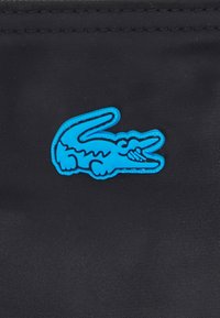 Le tissu noir présente un logo de crocodile bleu, souligné d'un contour bleu clair. Le design est plat et de style simpliste.