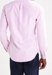 Koszula z długim rękawem w jasnoróżowym kolorze z białymi pionowymi paskami. Posiada zaokrąglony dół i mankiety zapinane na guziki. Materiał wydaje się gładki i miękki.