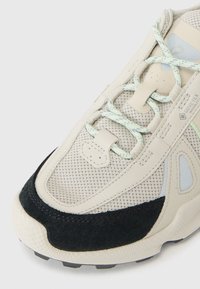 Beige Sneaker mit Mesh-Obermaterial, schwarzen Wildlederelementen und hellblauen Details. Verfügt über flache Schnürsenkel und eine strukturierte Sohle für besseren Halt.