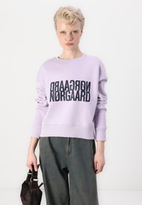 Violetinė džemperis su storu juodu užrašu "Nørgaard". Apvalus kaklas, ilgomis rankovėmis ir plačiais rankogaliais. Sukombinuota su laisvais, tamsiai žaliais kelnes.