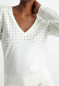 White knit sweater with a V-neckline, featuring scattered metallic square studs across the chest and long sleeves with ribbed cuffs.