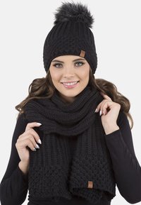 Cappello di maglia nero con pompon e sciarpa abbinata, caratterizzati da un motivo testurizzato e un accento in pelle. Entrambi gli articoli sono morbidi e caldi.