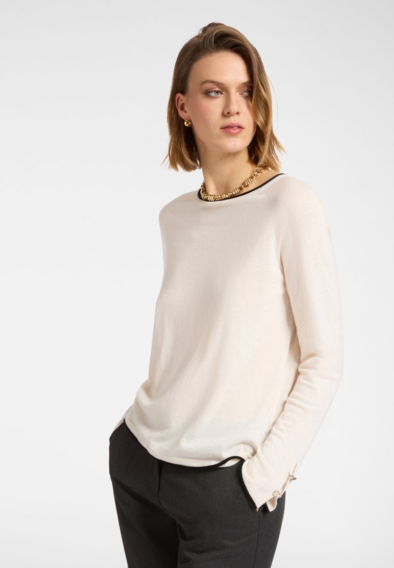 Helle beige Bluse mit langen Ärmeln aus weichem Stoff, mit einem runden Halsausschnitt mit schwarzem Besatz und Knopfdetails an den Manschetten.