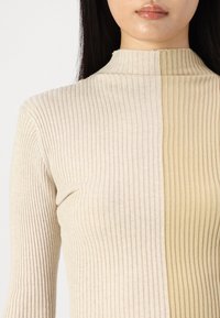 Bež ribani pulover s turtleneck ovratom, koji prikazuje vertikalne pruge s glatkom teksturom; podijeljen na svijetle bež i tamnije bež dijelove.